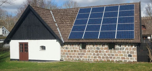 Ein Haus mit einer Fassade aus weißen und steinernen Mauern, braunen Doppeltüren und kleinen Fenstern. Auf dem braunen Ziegeldach sind mehrere blaue Solarmodule installiert.
