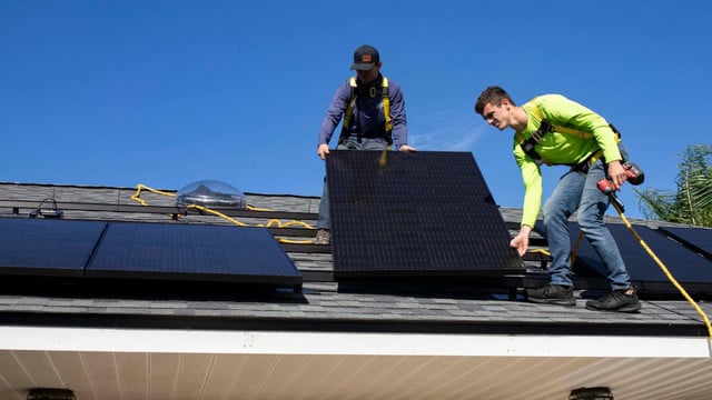Zwei Männer installieren Solarpaneele auf einem Hausdach. Beide tragen Sicherheitsgurte, einer hält ein Solarpanel, der andere einen Akkuschrauber.