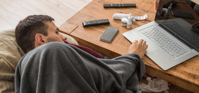 Ein Mann liegt mit einer grauen Decke zugedeckt auf einem Sofa und benutzt einen silbernen Laptop auf einem Holztisch. Auf dem Tisch liegen zwei Fernbedienungen, ein Smartphone, mehrere Medikamentenbehälter und eine zerknüllte Tablettenverpackung. Unter dem Tisch sind gebrauchte Papiertaschentücher sichtbar.