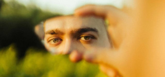Nahaufnahme von grünen Augen, die in einem kleinen, zerbrochenen Spiegel reflektiert werden, der von einer Hand gehalten wird. Der Hintergrund ist unscharf und zeigt grüne Pflanzen.