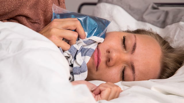 Warum werden Zahnschmerzen nachts oft schlimmer? Frau liegt im Bett und hält eine blaue Kältekompresse mit einem Tuch an ihre Wange, die Augen sind geschlossen.