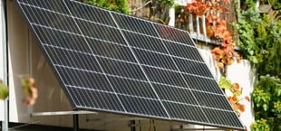 Zwei Solarpaneele sind an ein Balkongeländer montiert, umgeben von grünen und herbstlich gefärbten Pflanzen.