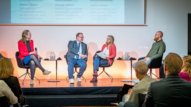 FairFinanceWeek_OSC2855_1920x1080px Podiumsdiskussion mit vier Personen auf Stühlen, zwei kleine Tische mit Wasserflaschen und Gläsern, Publikum im Vordergrund, Präsentation mit Text im Hintergrund.
