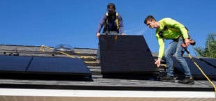 Zwei Männer installieren Solarpaneele auf einem Hausdach. Beide tragen Sicherheitsgurte, einer hält ein Solarpanel, der andere einen Akkuschrauber.