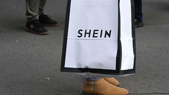 Frankreich weist Shein in die Schranken – wie reagiert Deutschland? Eine Einkaufstüte mit dem Aufdruck "SHEIN" in schwarzer Schrift auf weißem Hintergrund mit schwarzem Rand, gehalten von einer Person mit braunen Wildlederstiefeln auf grauem Asphalt. Im Hintergrund sind weitere Personen mit dunklen Schuhen zu sehen.