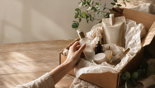beauty-online-bestellung-hero Hand, die eine durchsichtige Kosmetikflasche mit Pumpspender aus einem offenen Karton mit verschiedenen Kosmetikprodukten hält, darunter ein braunes Pipettenfläschchen, ein Cremebehälter, ein Sprayflakon und eine Nachfüllpackung, alles auf einem Holztisch mit weichem Tageslicht und grünen Blättern im Hintergrund.