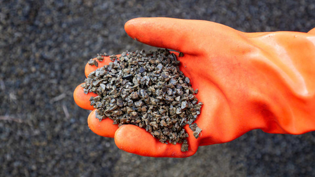 Streusalz-Alternativen: Was Sand, Kies und Asche besser machen - und wann du aufpassen musst Hand mit orangefarbener Schutzhandschuh, die eine Menge kleiner, grauer Kieselsteine hält, vor einem unscharfen Hintergrund aus ähnlichem Material.