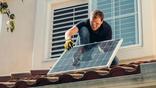 Photovoltaik: Der richtige Neigungswinkel steigert den Ertrag Ein weißer Mann mit Schutzbrille und Arbeitshandschuhen installiert ein Solarpanel auf einem Dach mit roten Ziegeln .