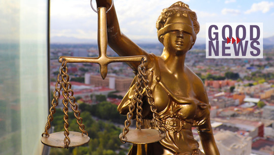 gerechtigkeit-pb-2606 Goldene Justitia-Statue mit Waage vor unscharfem Stadtpanorama, daneben Schriftzug "Good News" mit roten Pfeilen.