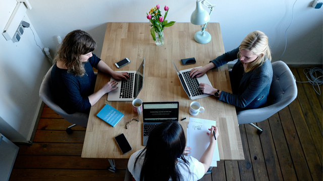 Drei Frauen sitzen an einem Holztisch, zwei arbeiten an Laptops, eine schreibt auf Papier. Auf dem Tisch stehen zwei Kaffeetassen, ein Blumenstrauß, eine Schreibtischlampe, mehrere Smartphones, eine Brille und ein Buch. Der Raum hat Holzboden und weiße Wände.