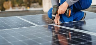 Eine Person in blauer Arbeitskleidung und grauem Pullover montiert Solarpaneele auf einem Dach und benutzt dabei einen Akkuschrauber.