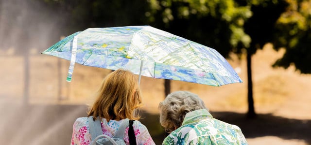 Bericht: Millionen Menschen sterben wegen Klimakrise – eine Maßnahme rettet etwa 160.000 Leben pro Jahr (Foto: Rolf Vennenbernd/dpa) Zwei Frauen von hinten, eine mit roten Haaren und Rucksack, die andere mit grauen Haaren, die gemeinsam einen bunten Regenschirm mit floralen Mustern halten. Im Hintergrund unscharfe Bäume und ein sonniger Tag.