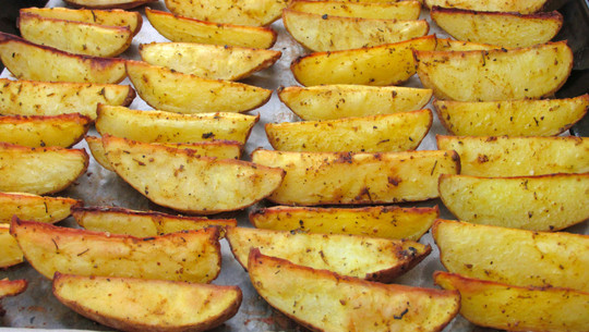 Jeden Tag Kartoffeln essen: Das sind die Folgen Mehrere goldgelbe, gewürzte Kartoffelspalten liegen gleichmäßig auf einem Backblech mit Backpapier, leicht knusprig gebacken.