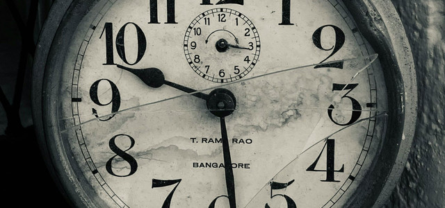 Alte, runde Wanduhr mit großen schwarzen Zahlen auf weißem Zifferblatt, das durch Risse und Flecken gezeichnet ist. Die Uhr zeigt die Zeit 10:29, mit einem kleineren Zifferblatt oben für Sekunden. Auf dem Zifferblatt steht "T. RAM RAO" und "BANGALORE".