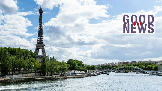 "Eine Art Wunderlösung": Paris setzt auf Alternative zu Klimaanlagen Eiffelturm in Paris, umgeben von Bäumen, mit der Seine im Vordergrund und einer Brücke im Hintergrund, unter einem bewölkten Himmel. Rechts im Bild steht der Schriftzug „GOOD NEWS“ in großen lila Buchstaben mit drei roten Pfeilen dazwischen.