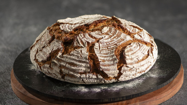 Bäckerverband: Das ist das Brot des Jahres 2026 Rundes, rustikales Sauerteigbrot mit knuspriger, rissiger Kruste und Mehlbestäubung, liegend auf einer schwarzen runden Platte mit Holzunterlage.