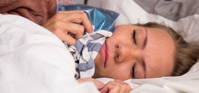 Frau liegt im Bett und hält eine blaue Kältekompresse mit einem Tuch an ihre Wange, die Augen sind geschlossen.