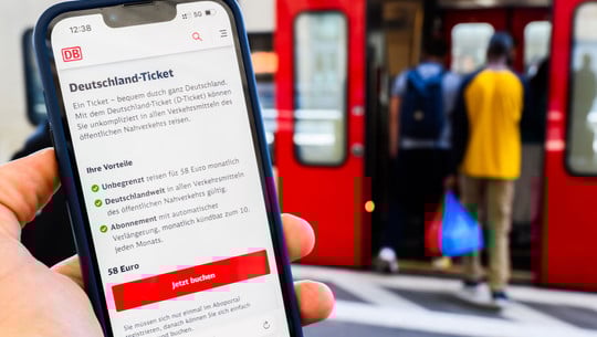 Gasheizung, Deutschlandticket, Schweinefleisch: Das ändert sich 2026 Hand, die ein Smartphone mit der Buchungsseite für das Deutschland-Ticket der Deutschen Bahn zeigt, im Hintergrund steigen zwei Personen in eine rote Bahn ein.