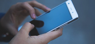 Hände, die ein weißes Smartphone mit schwarzem Bildschirm halten und bedienen, vor unscharfem grauem Hintergrund.