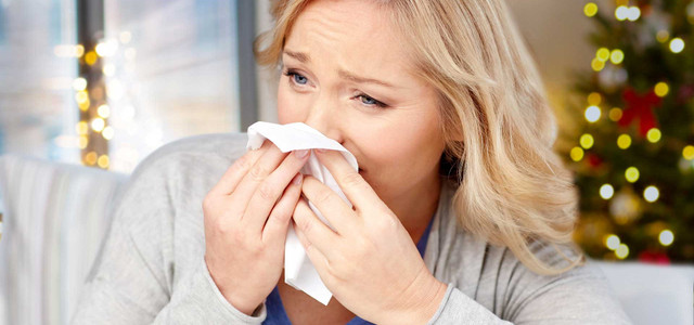 Krank an Weihnachten: Was tun, wenn die Arztpraxis geschlossen ist? (Foto: Colourbox/ 34963881) Frau mit blonden Haaren, die sich mit einem Taschentuch die Nase putzt, im Hintergrund unscharfer Weihnachtsbaum mit Lichtern.