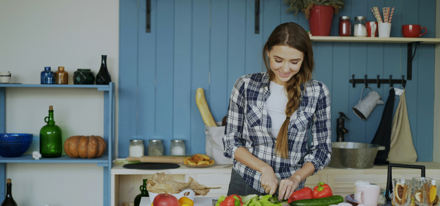 Planetary Health Diet: So gelingt der Einstieg in eine bewusste und nachhaltige Ernährung (CC0 Vitaly Gariev / Unsplash) Eine junge Frau mit geflochtener Haarsträhne schneidet in einer Küche Paprika und Salat auf einem Tisch. Im Hintergrund sind Regale mit verschiedenen Küchenutensilien, Gläsern und einem Kürbis zu sehen. Auf dem Tisch liegen außerdem ein Apfel, Zitronen, eine Gurke, Tassen und Gläser mit getrockneten Früchten. Die Wand hinter ihr ist blau vertäfelt.