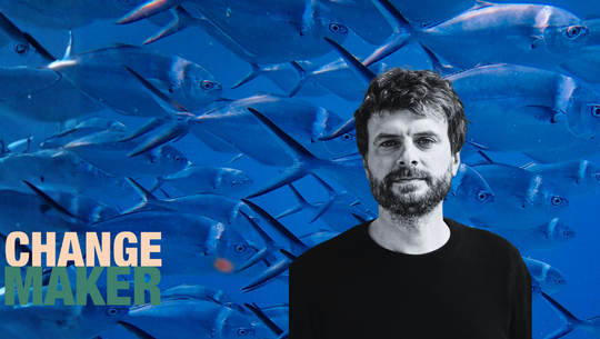 Kopie von Podcast Changemaker - Artikelbild(5) Ein Mann mit dunklem, lockigem Haar und Bart trägt ein schwarzes Oberteil. Er steht vor einem Hintergrund mit vielen blauen Fischen, die dicht beieinander schwimmen. Links unten steht der Text „CHANGE MAKER“ in großen Buchstaben, wobei „CHANGE“ in hellbeige und „MAKER“ in dunkelgrün geschrieben ist.