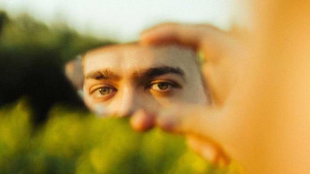 Wie Widersprüche aushalten – auch in uns selbst? Therapeutin gibt Tipps und erklärt, warum es wichtig ist Nahaufnahme von grünen Augen, die in einem kleinen, zerbrochenen Spiegel reflektiert werden, der von einer Hand gehalten wird. Der Hintergrund ist unscharf und zeigt grüne Pflanzen.