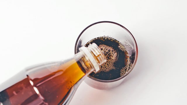 Jeden Tag Softdrinks: Die Folgen Eine braune Flasche gießt eine dunkle, kohlensäurehaltige Flüssigkeit in ein durchsichtiges Glas auf weißem Hintergrund.