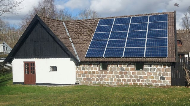 Photovoltaikanlagen im Denkmalschutz Ein Haus mit einer Fassade aus weißen und steinernen Mauern, braunen Doppeltüren und kleinen Fenstern. Auf dem braunen Ziegeldach sind mehrere blaue Solarmodule installiert.