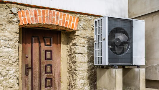 Langzeitstudie: Wärmepumpen sind auch im Altbau effizient und klimaschonend Außeneinheit einer Wärmepumpe, montiert auf zwei Betonblöcken neben einer alten Steinmauer mit einer braunen Holztür und einem Bogen aus roten Ziegelsteinen darüber.