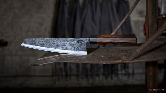 header-01 Küchenmesser mit einer grob strukturierten, dunklen Klinge und einem glatten, dunkelbraunen Holzgriff, liegend auf einer rostigen Metallfläche vor unscharfem, dunklem Hintergrund. Auf der Klinge steht der Schriftzug "BLACK CHILI".