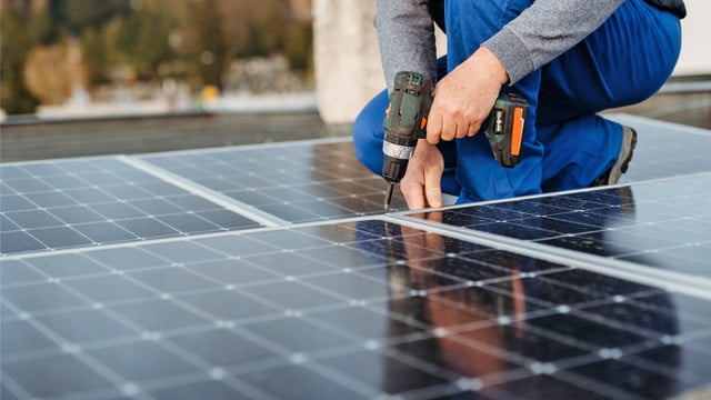 Handwerker installiert Photovoltaik-Module auf einem Dach Eine Person in blauer Arbeitskleidung und grauem Pullover montiert Solarpaneele auf einem Dach und benutzt dabei einen Akkuschrauber.