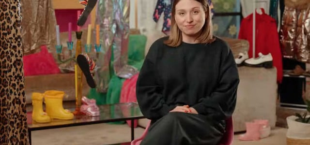 Bianca Heinicke sitzt auf einem pinken Stuhl. Sie trägt einen schwarzen Pullover und einen schwarzen Rock. Im Hintergrund sind bunte Kleidungsstücke, darunter ein Leopardenmuster, gelbe Gummistiefel, ein roter Mantel, weiße Schuhe und pinke Stiefel zu sehen. Außerdem sind ein Kerzenhalter mit bunten Kerzen und ein Tisch mit roten Tomaten darauf zu erkennen.