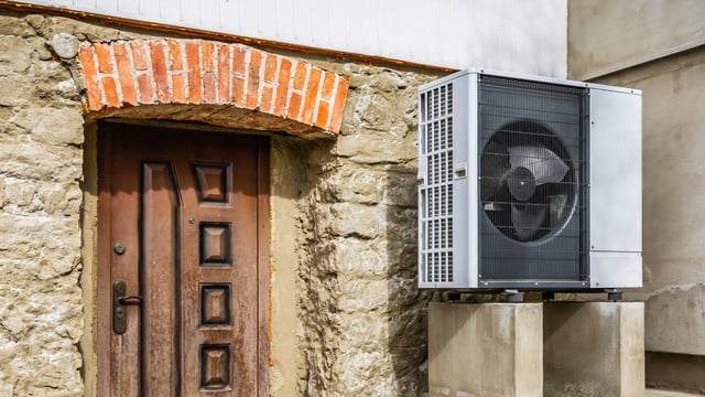 Langzeitstudie: Wärmepumpen sind auch im Altbau effizient und klimaschonend Außeneinheit einer Wärmepumpe, montiert auf zwei Betonblöcken neben einer alten Steinmauer mit einer braunen Holztür und einem Bogen aus roten Ziegelsteinen darüber.