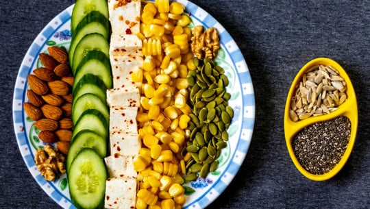 Nurtrient Sequencing Richtige Reihenfolge Lebensmittel Ein Teller mit in Reihen angeordneten Mandeln, Gurkenscheiben, gewürztem Tofu, Maiskörnern, Walnüssen und Kürbiskernen. Daneben eine gelbe Schale mit Sonnenblumenkernen und Chiasamen.
