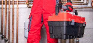 Heizungsmonteur in roter Arbeitskleidung hält einen schwarzen und roten Werkzeugkasten vor einer Wand mit Heizungsrohren