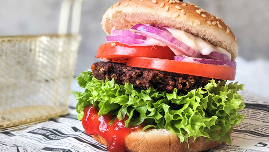 EU vertagt Entscheidung: Bleibt der Veggie-Burger ein Burger? Hamburger mit veganem Patty und Sesambrötchen, grünem Salat, Tomatenscheiben, roten Zwiebeln, einer dunklen, knusprigen Pattieschicht, Mayonnaise und Ketchup, serviert auf Zeitungspapier.