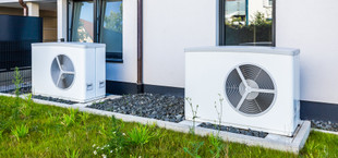 Zwei weiße Wärmepumpen stehen auf Betonplatten vor einem modernen Haus mit weißen Wänden und schwarzen Fensterrahmen.