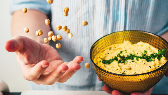 Wie eine Ernährungsexpertin es schafft, jeden Tag ausreichend Protein zu essen Eine Person hält eine Schüssel mit Hummus. Mit der anderen Hand wirft sie Kichererbsen in die Luft.