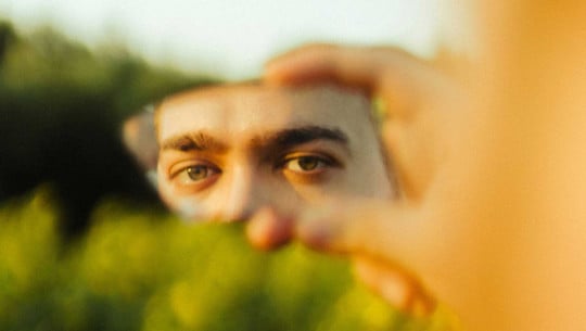 Wie Widersprüche aushalten – auch in uns selbst? Therapeutin gibt Tipps und erklärt, warum es wichtig ist Nahaufnahme von grünen Augen, die in einem kleinen, zerbrochenen Spiegel reflektiert werden, der von einer Hand gehalten wird. Der Hintergrund ist unscharf und zeigt grüne Pflanzen.
