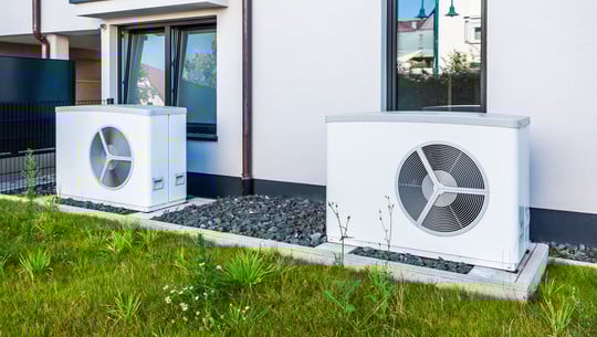Wärmepumpen-Förderung: Neue Bedingungen ab 2026 Zwei weiße Wärmepumpen stehen auf Betonplatten vor einem modernen Haus mit weißen Wänden und schwarzen Fensterrahmen.