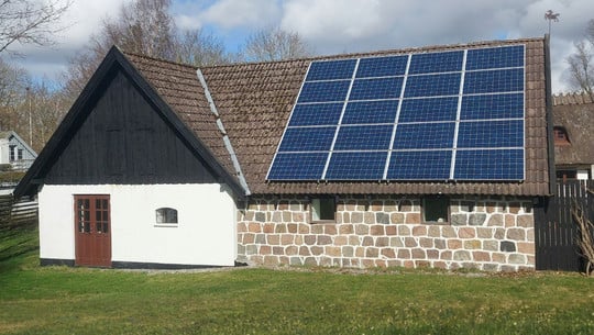 Photovoltaikanlagen im Denkmalschutz Ein Haus mit einer Fassade aus weißen und steinernen Mauern, braunen Doppeltüren und kleinen Fenstern. Auf dem braunen Ziegeldach sind mehrere blaue Solarmodule installiert.