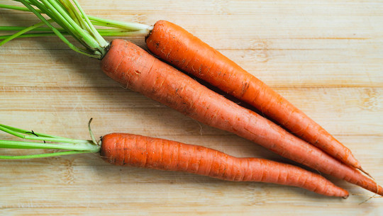 Jeden Tag Karotten essen: Das sind die Folgen Drei frische, noch leicht erdige Karotten mit grünen Stielen liegen auf einem hellen Holzbrett.