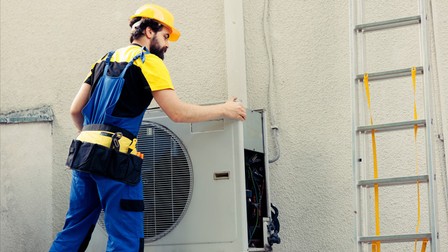 Ein Handwerker mit gelbem Schutzhelm, gelb-schwarzem T-Shirt und blauer Arbeitslatzhose öffnet das Gehäuse einer Wärmepumpe an einer Außenwand.