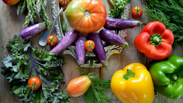 nadine-primeau-wpoKpJqOsKE-unsplash Verschiedene frische Gemüsesorten auf einem Holztisch, darunter lila Auberginen, rote, gelbe und grüne Paprika, grüne Spargelstangen, Tomaten in verschiedenen Größen, grüner Grünkohl und Dill. Die Auberginen und eine große Tomate liegen auf einem silbernen Tablett.