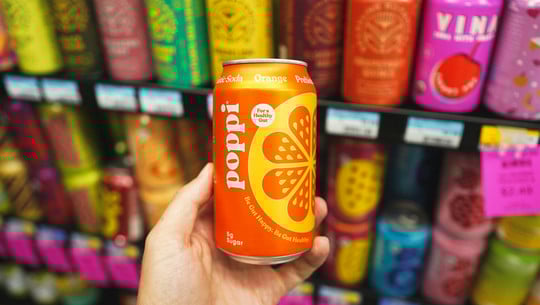 Functional Drinks Hand hält einen Functional Drink: eine orangefarbene Dose "poppi" mit Orangen-Geschmack, im Hintergrund unscharfe bunte Getränkedosen in einem Kühlschrank.