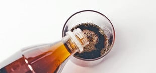 Eine braune Flasche gießt eine dunkle, kohlensäurehaltige Flüssigkeit in ein durchsichtiges Glas auf weißem Hintergrund.