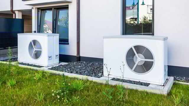 Wärmepumpen-Förderung: Neue Bedingungen ab 2026 Zwei weiße Wärmepumpen stehen auf Betonplatten vor einem modernen Haus mit weißen Wänden und schwarzen Fensterrahmen.