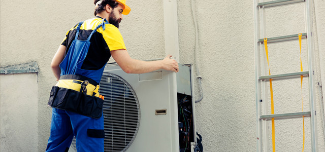 Ein Handwerker mit gelbem Schutzhelm, gelb-schwarzem T-Shirt und blauer Arbeitslatzhose öffnet das Gehäuse einer Wärmepumpe an einer Außenwand.