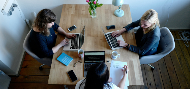 Drei Frauen sitzen an einem Holztisch, zwei arbeiten an Laptops, eine schreibt auf Papier. Auf dem Tisch stehen zwei Kaffeetassen, ein Blumenstrauß, eine Schreibtischlampe, mehrere Smartphones, eine Brille und ein Buch. Der Raum hat Holzboden und weiße Wände.